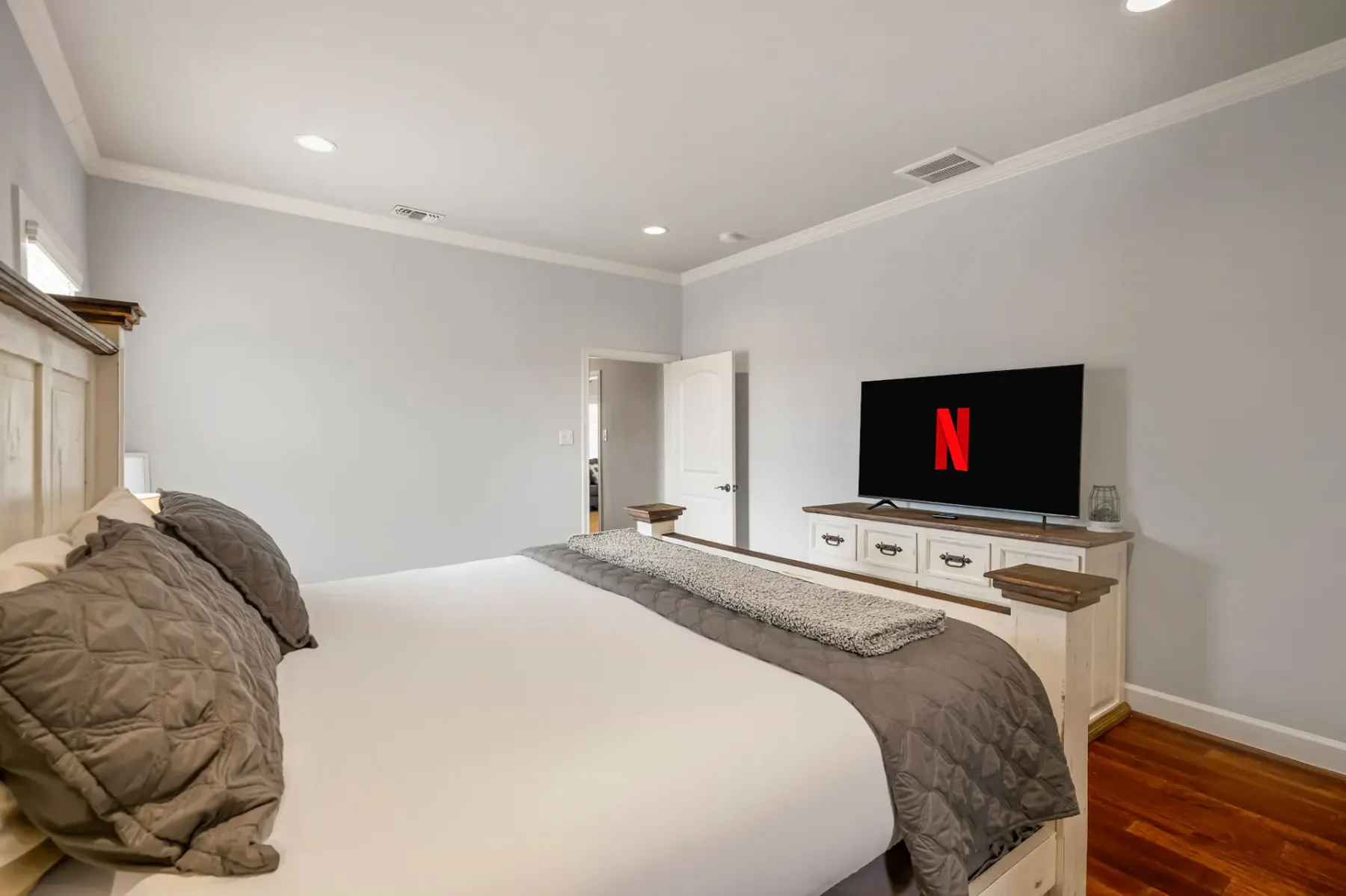 A modern bedroom with a large bed, gray and white bedding, a flat-screen TV on a dresser, and hardwood floors.
