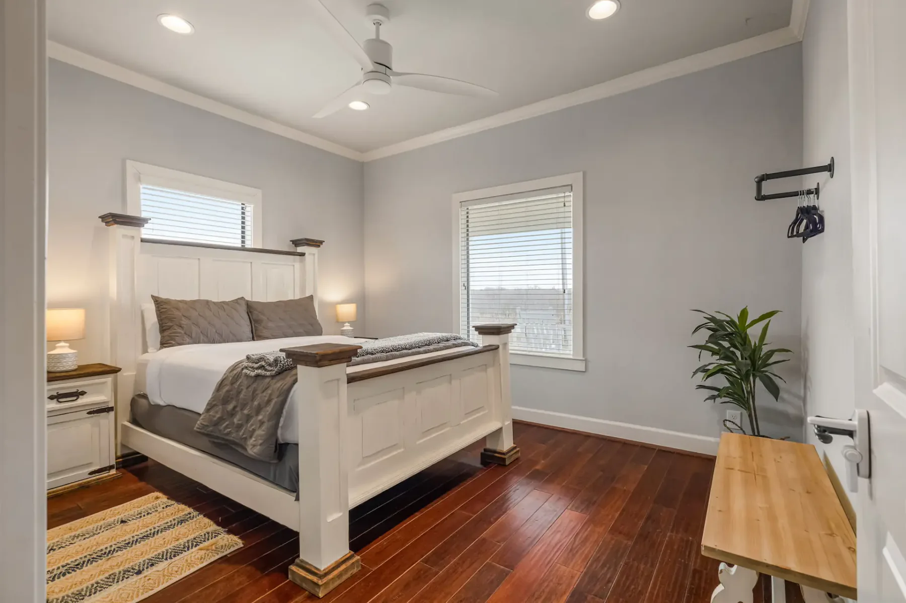 A tidy bedroom with a large white bed, wooden flooring, two windows, a ceiling fan, and a small plant in the corner. A bench sits at the foot of the bed.