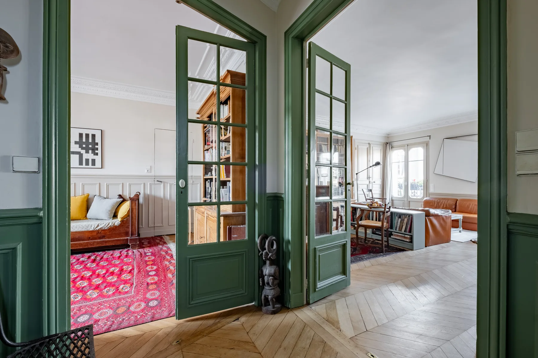 An interior room with open green-framed glass doors and bright living areas beyond.