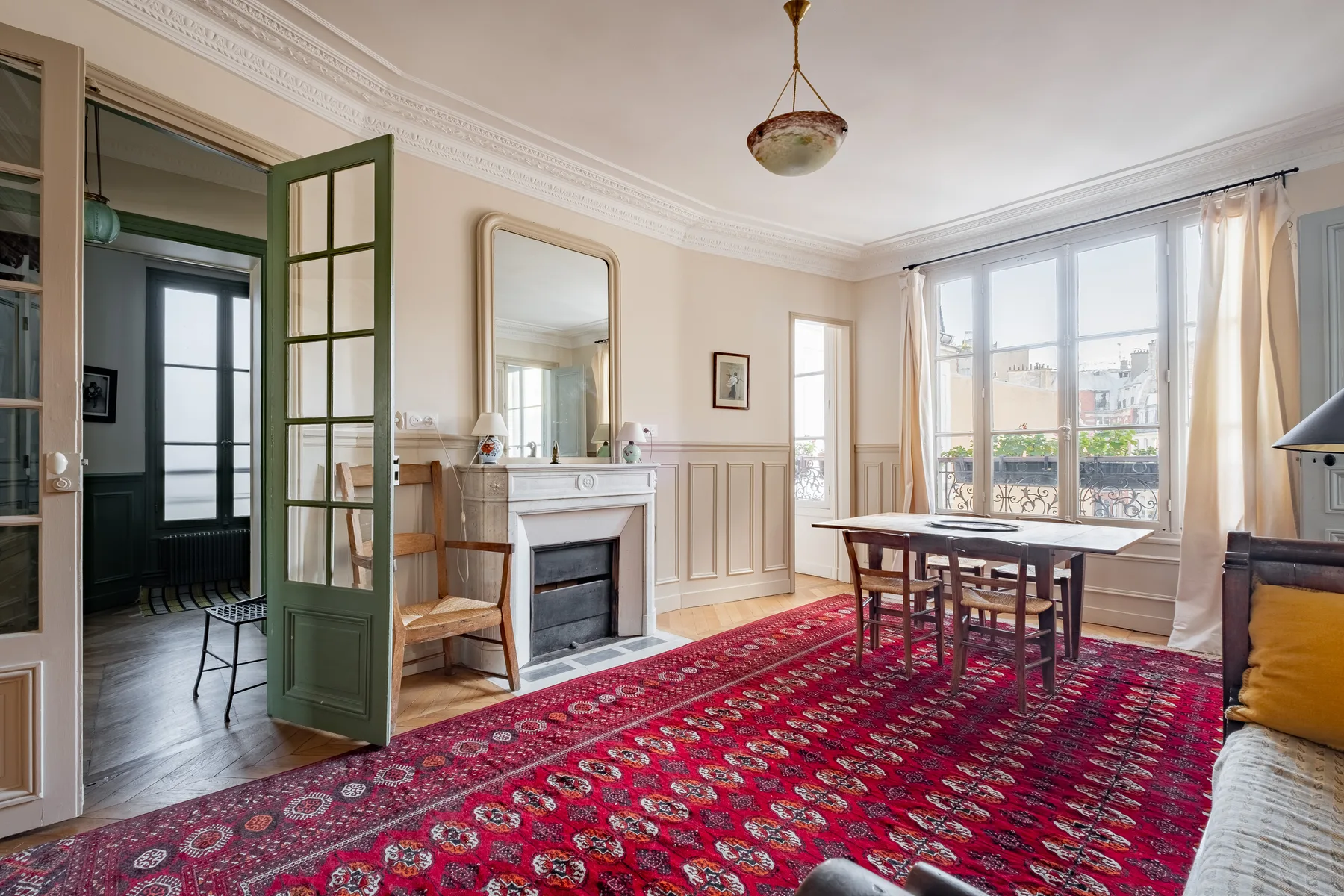 A bright dining room with a wooden table beside large windows and a red patterned rug.