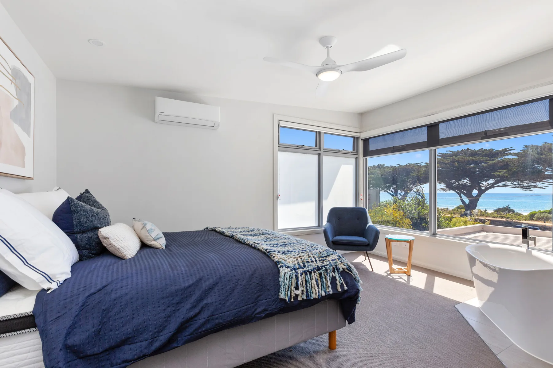 Un dormitorio luminoso con una cama, un sillón y grandes ventanas con vistas a la costa.