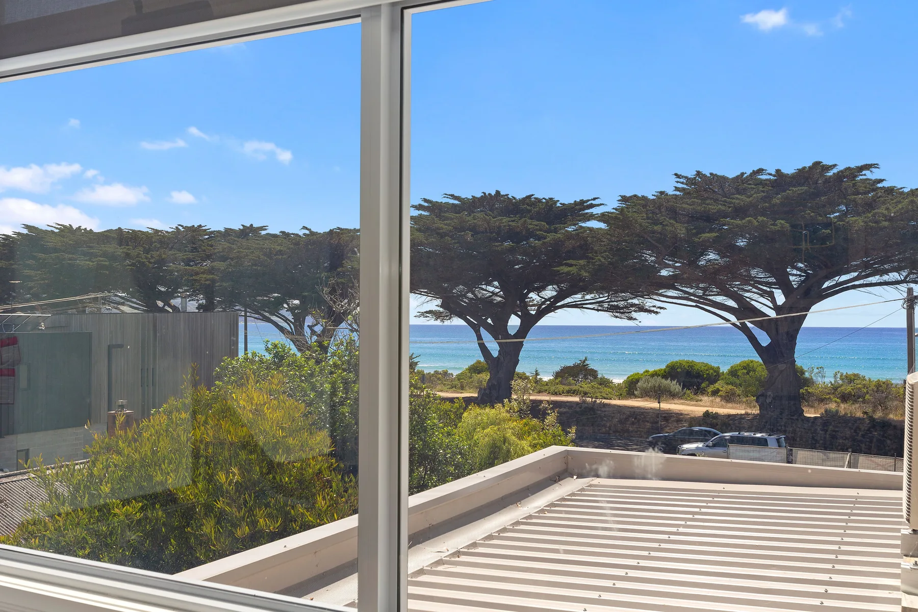 Una vista a través de grandes ventanas muestra árboles, el océano y una terraza soleada.