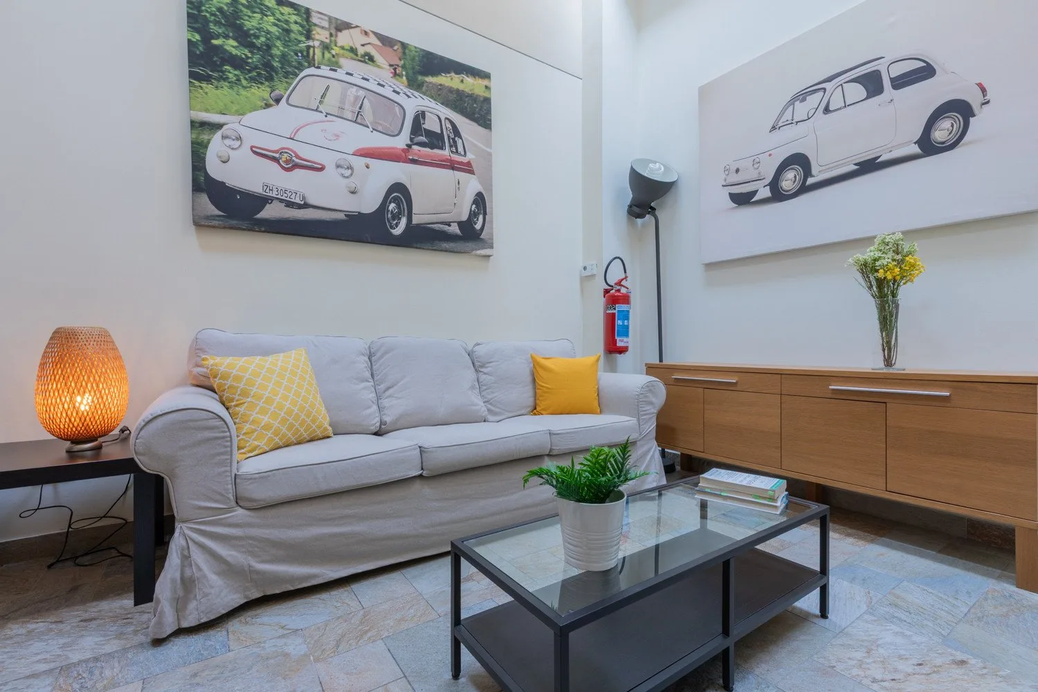 Una sala de estar con sofá, mesa de centro de vidrio e ilustraciones de autos clásicos.