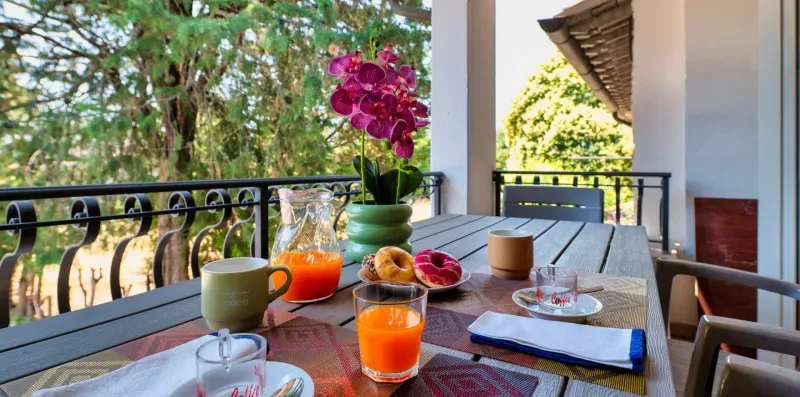 A balcony table is set for breakfast beside a railing and leafy trees.