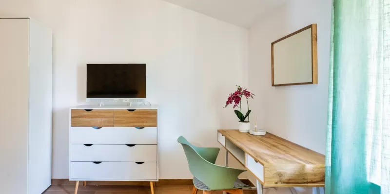A bright bedroom with a television on a dresser and a desk beside a window.