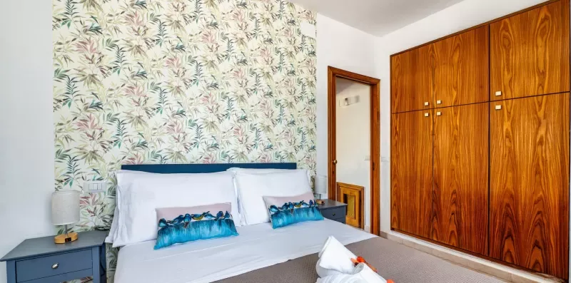 A bedroom with a bed, patterned wallpaper, and wooden wardrobes.