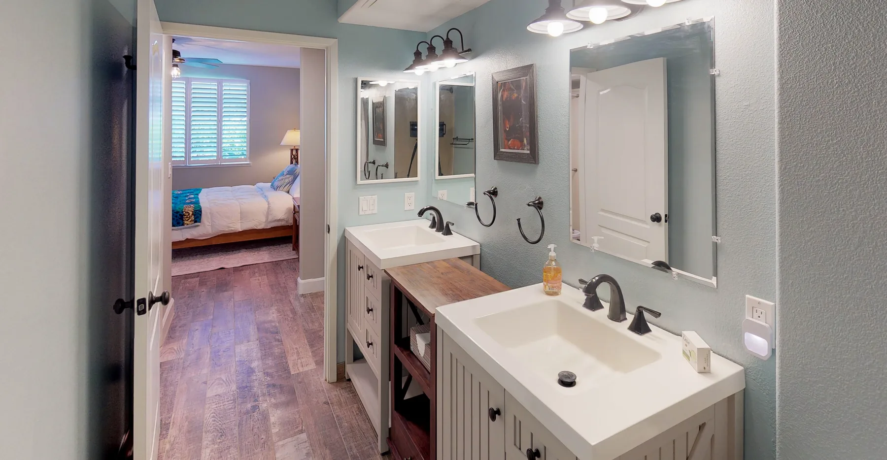 Un baño con dos lavabos y espejos, con un dormitorio visible a través de la puerta.