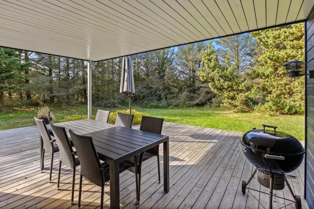 Una terraza cubierta de madera tiene una mesa de comedor, asientos y una parrilla con vistas a un jardín verde.