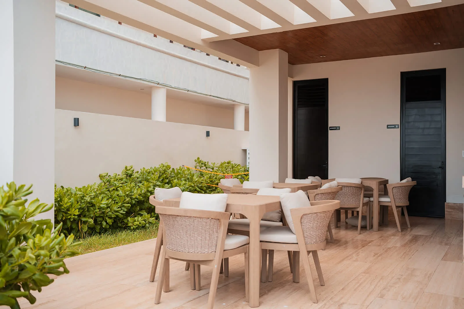 Una zona de comedor al aire libre con mesas y sillas junto a una hilera de plantas verdes.