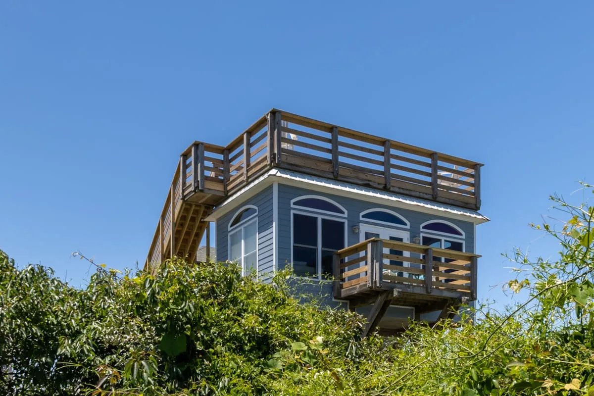Una casa azul con terrazas de madera se alza sobre vegetación frondosa bajo un cielo despejado.