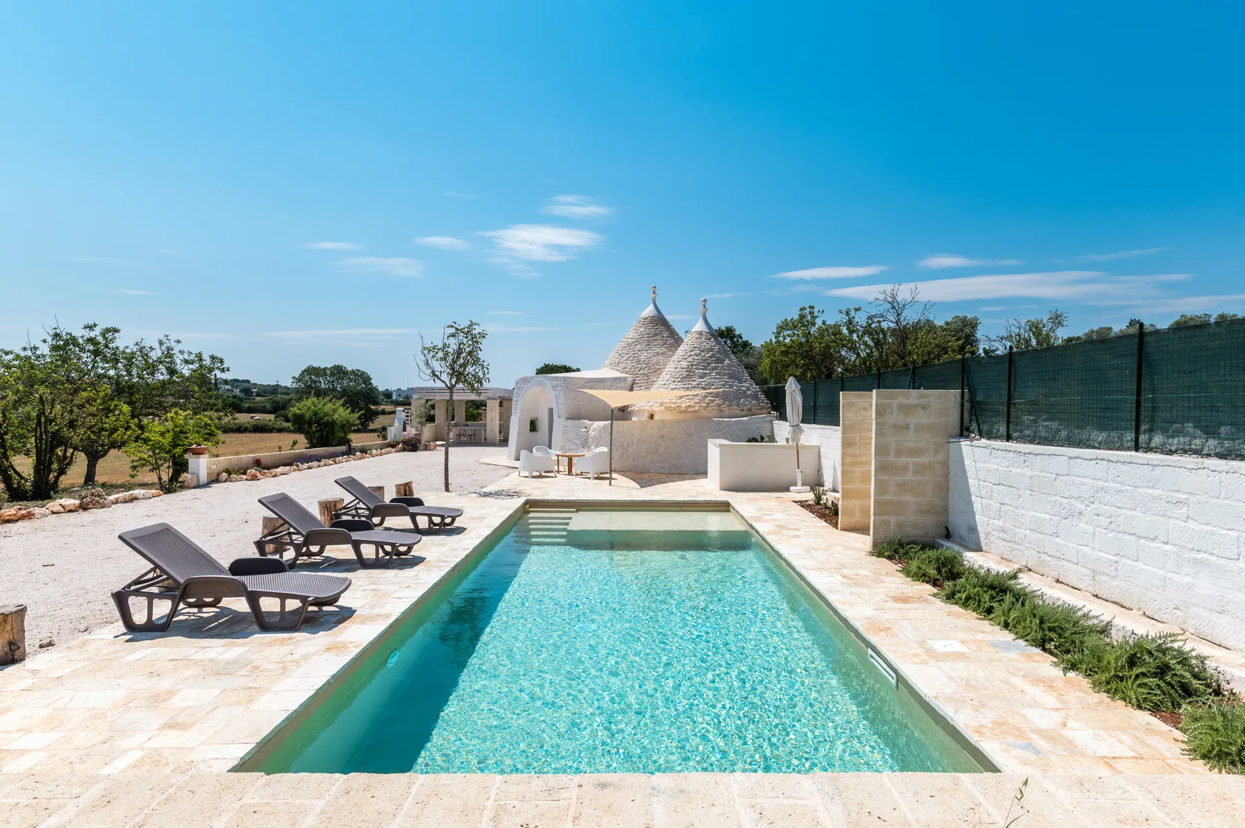 An outdoor swimming pool beside a paved patio with loungers and white stone buildings.