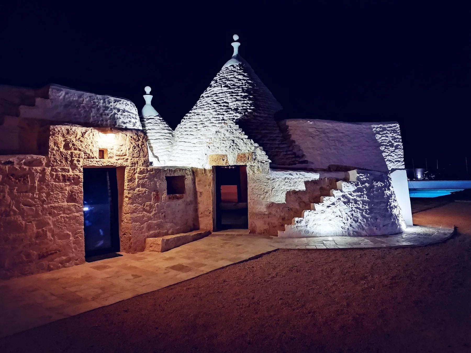 A stone building with conical roofs is illuminated at night beside a pool.