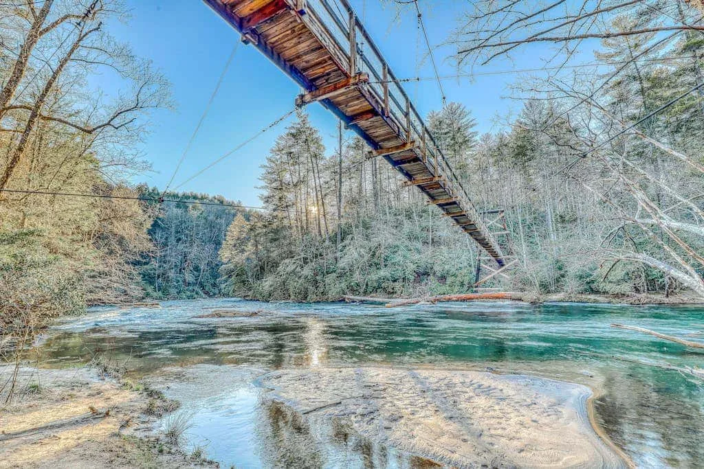 Un puente colgante cruza un río claro bordeado de árboles.