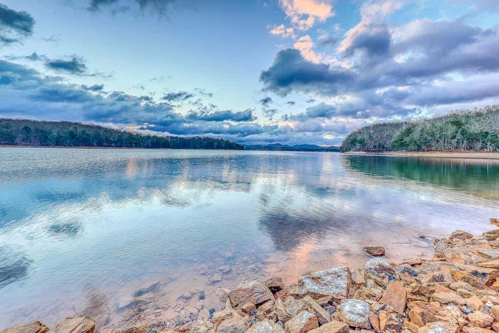 Un lago tranquilo refleja un cielo nublado, con rocas a lo largo de la orilla.