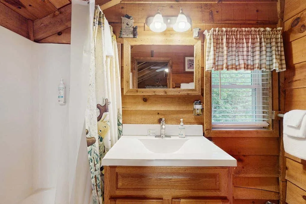 Baño revestido de madera con lavabo, espejo y ventana con cortina.