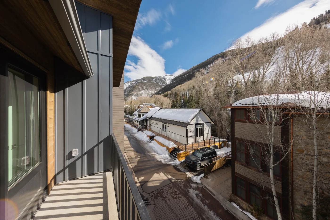 A balcony overlooks nearby buildings and a wooded mountain slope beneath a blue sky.