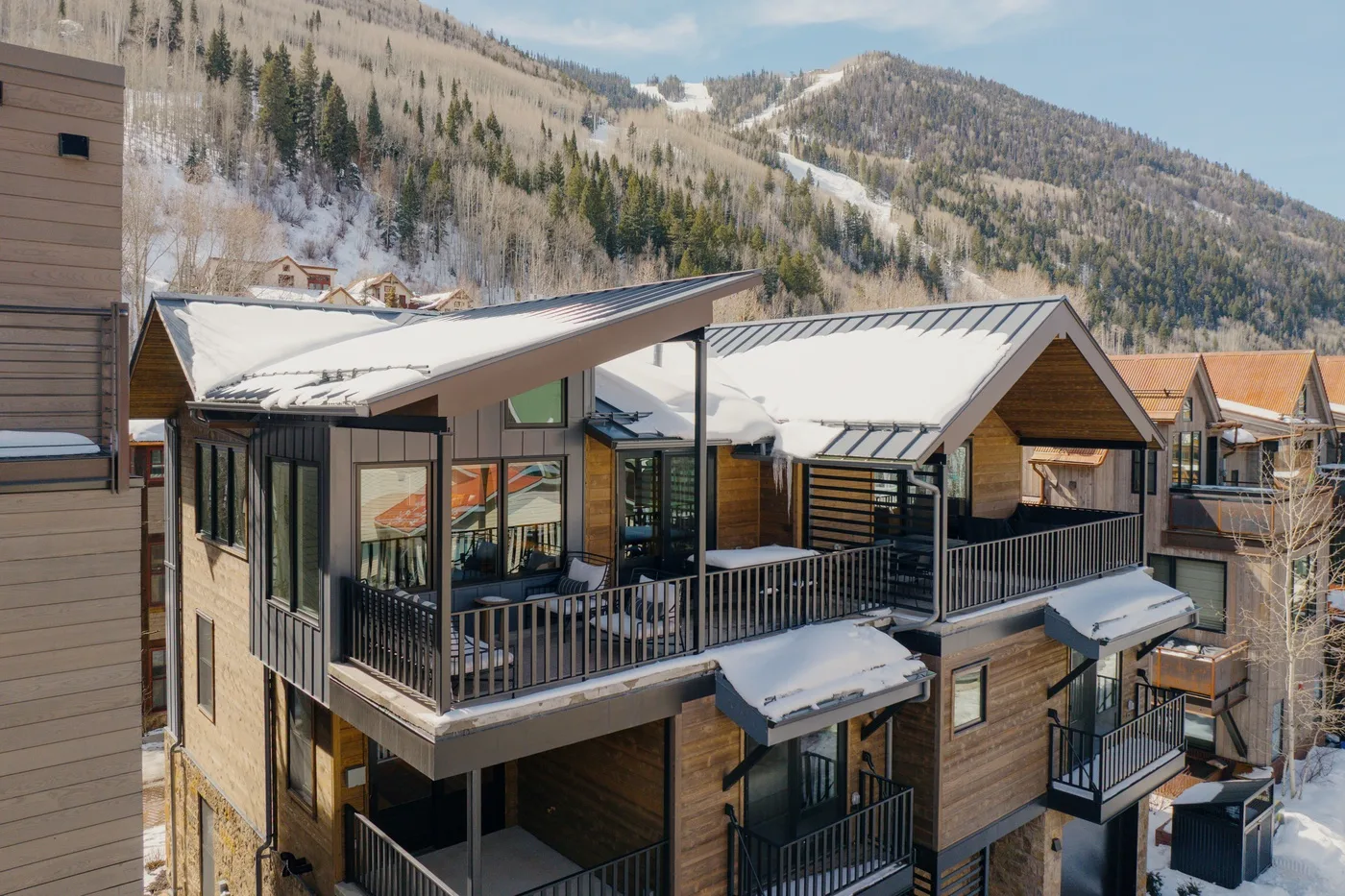 A modern mountain building with snow-covered roofs and balconies sits below wooded slopes.