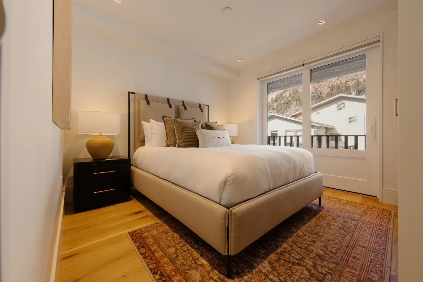 A bedroom with a bed, bedside table, and large window overlooking nearby buildings.