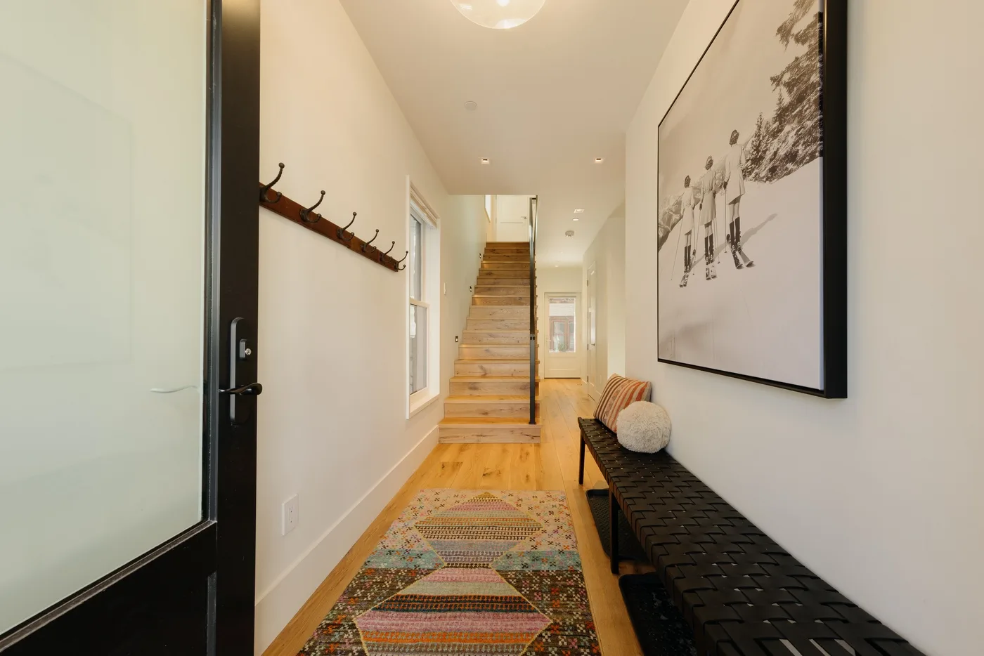 A narrow hallway has a staircase, wall hooks, and a long bench.