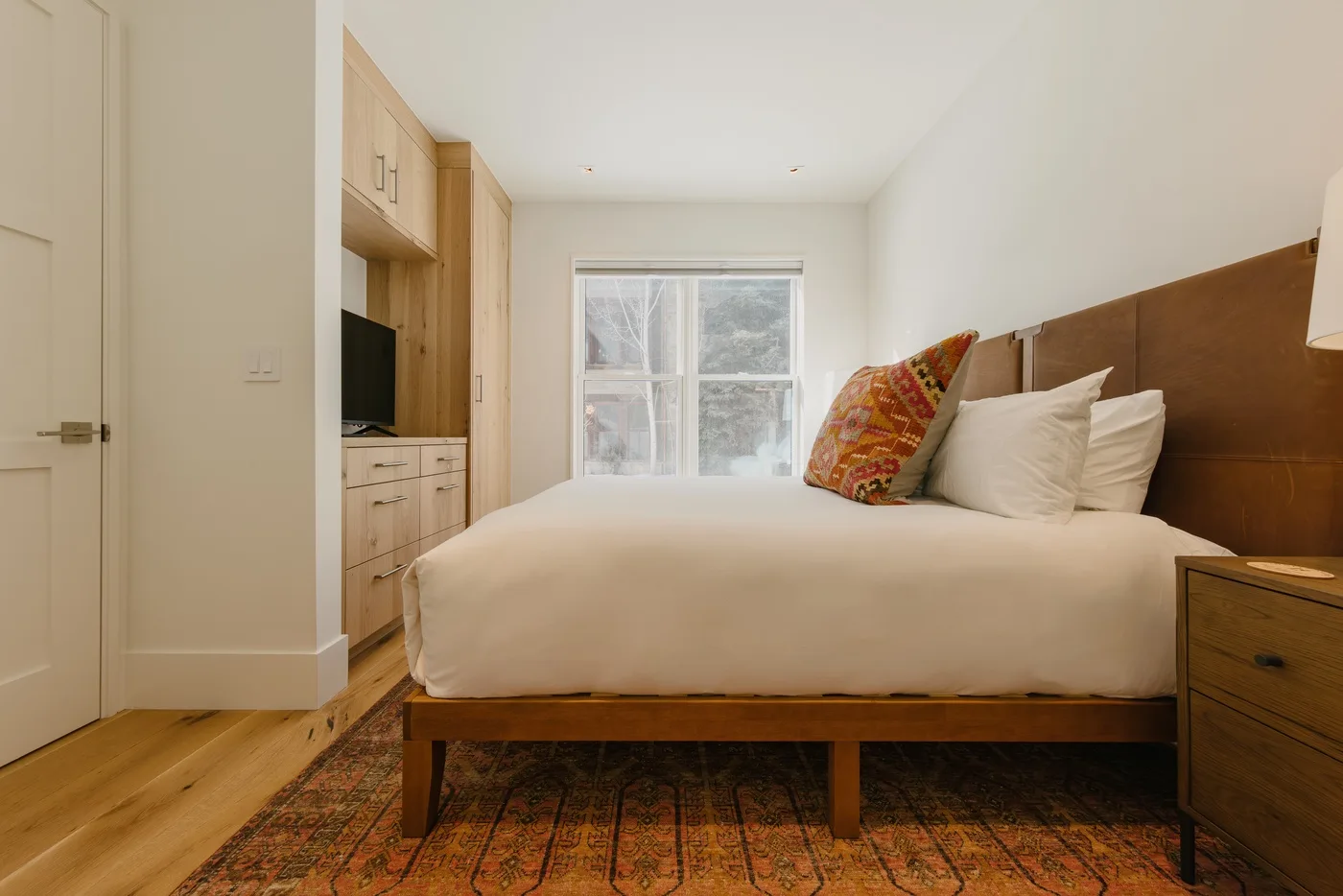 A bedroom with a neatly made bed, colorful accent pillow, and window.