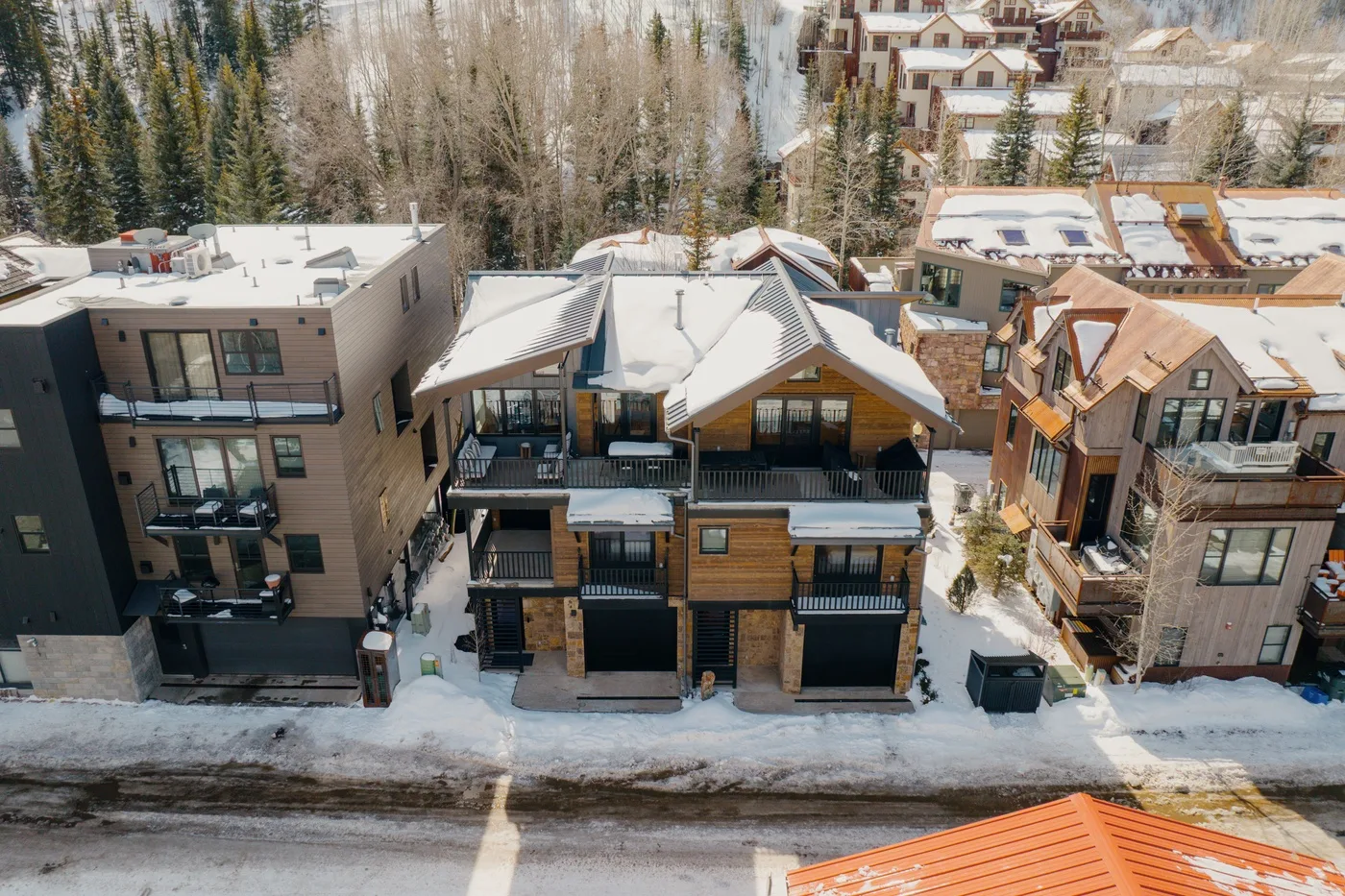 Aerial view of snow-covered buildings along a street, with evergreen trees behind them.