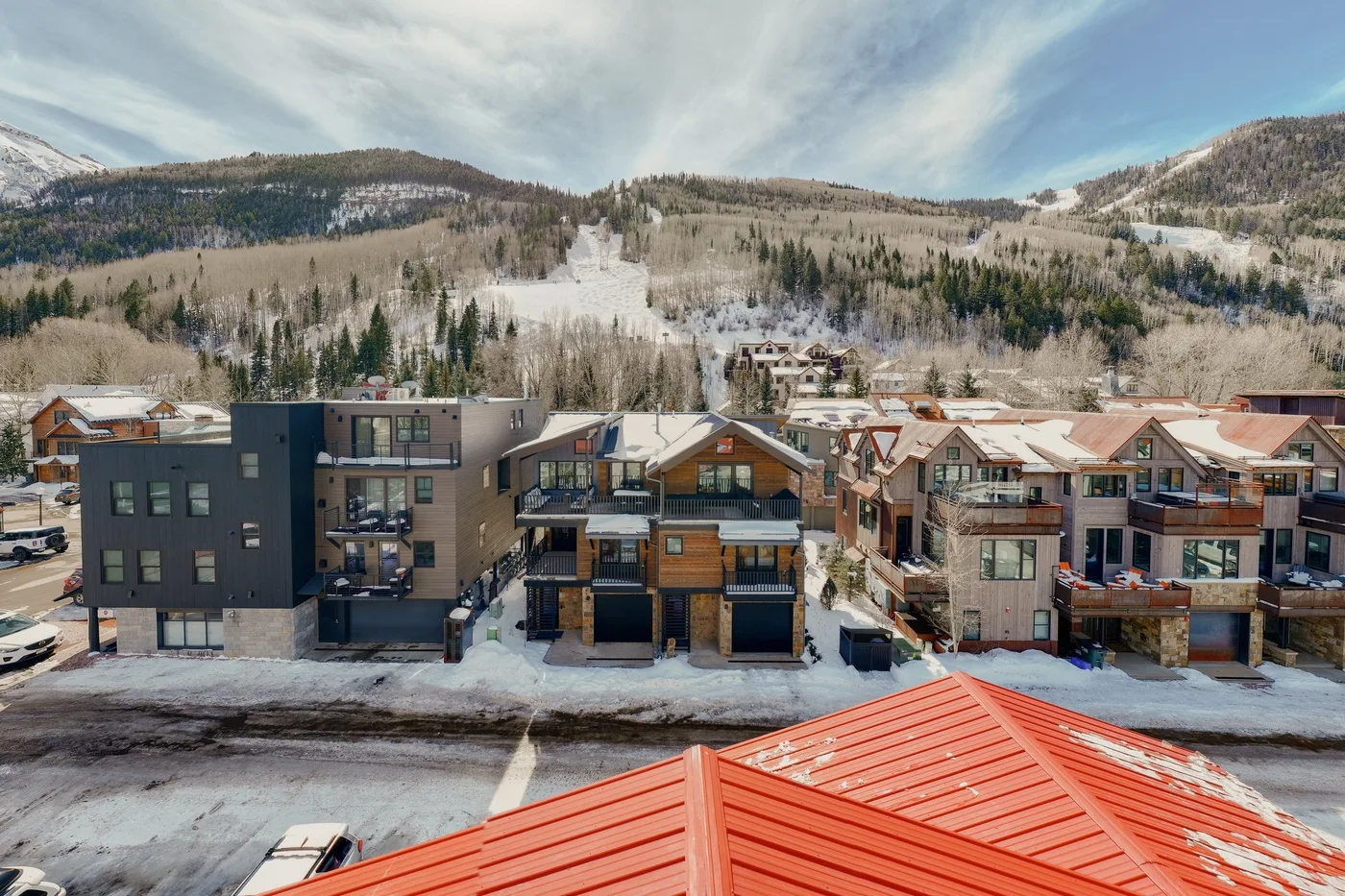 A row of mountain residences sits below a snowy hillside under a partly cloudy sky.