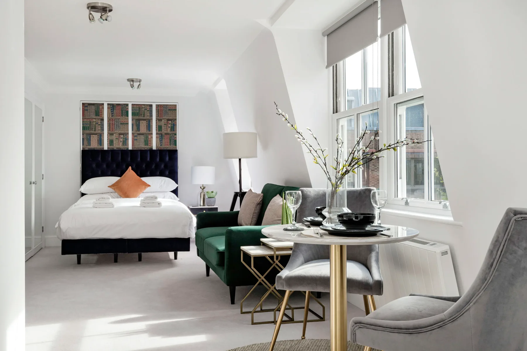A bright studio bedroom with a bed, green seating, and a dining table beside large windows.