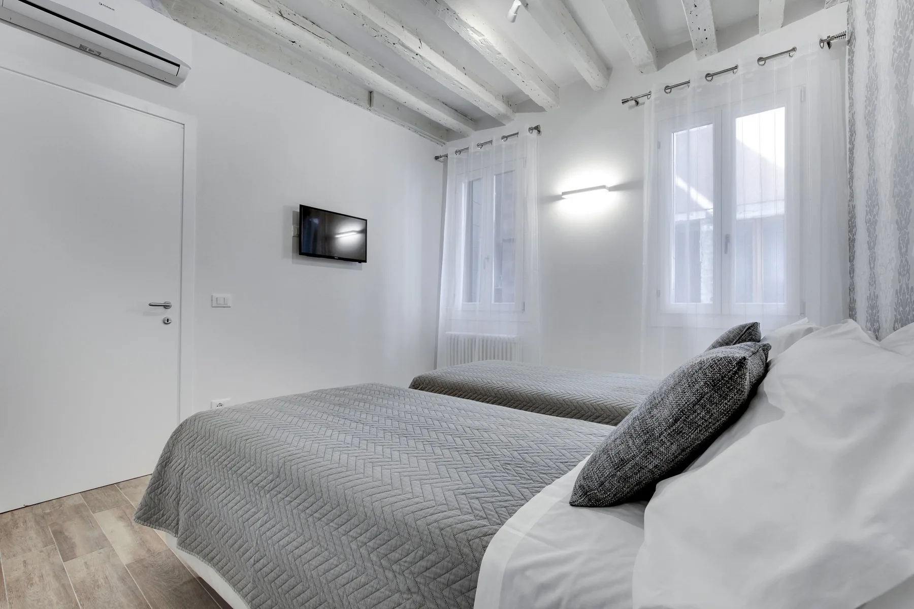 Un dormitorio luminoso con dos camas, ventanas con cortinas y un televisor en la pared.