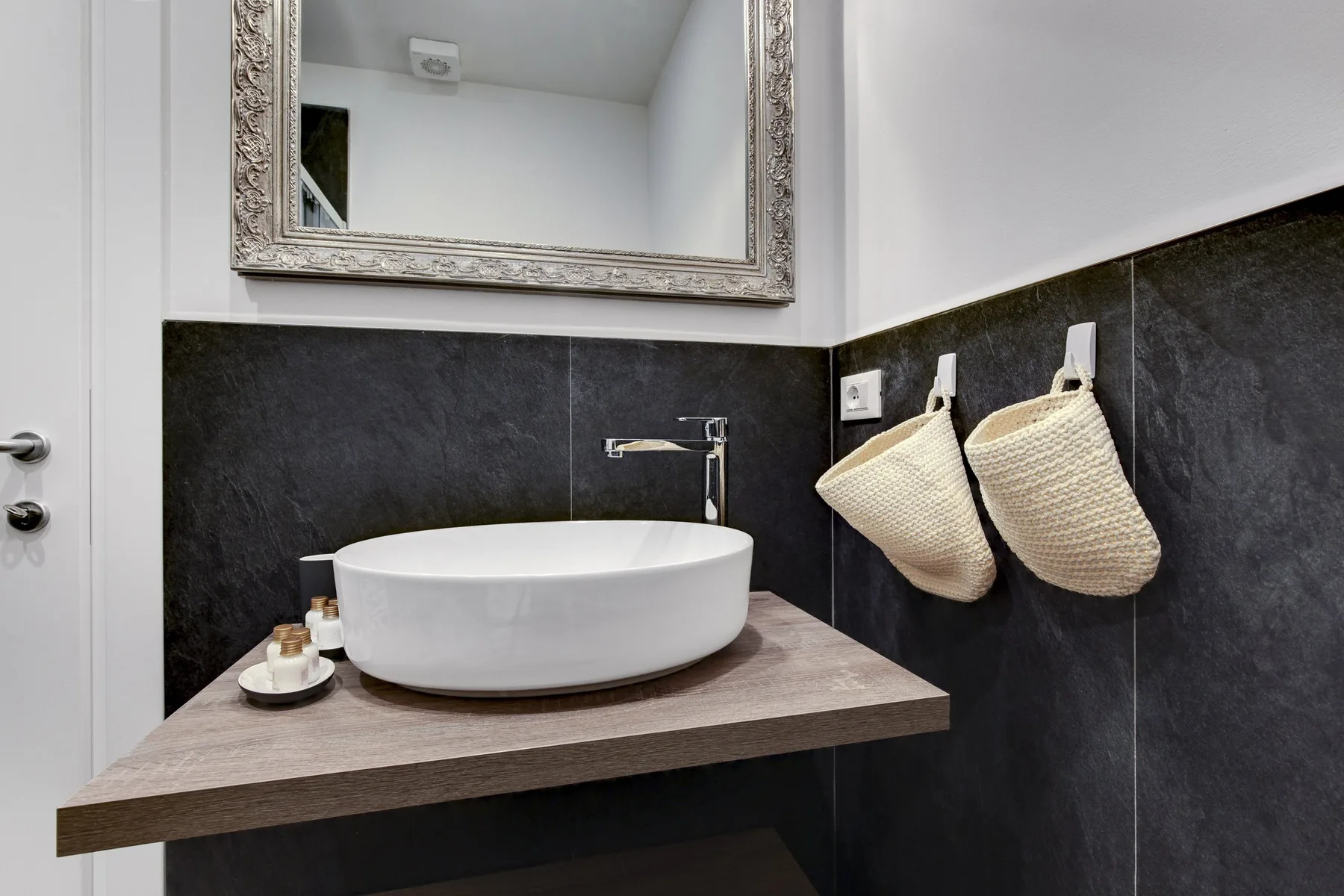 Un lavabo de baño con un lavabo blanco sobre encimera, paredes de azulejos oscuros y un espejo.