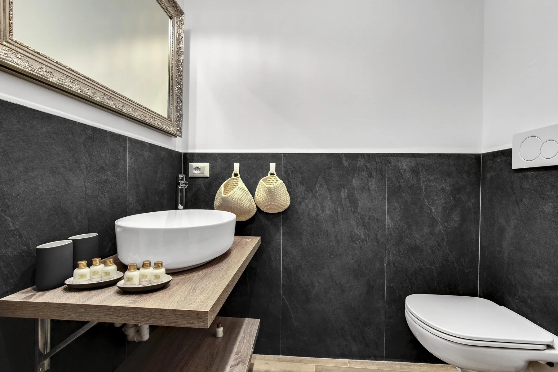 Un baño moderno con lavabo sobre encimera, azulejos oscuros y un inodoro suspendido.