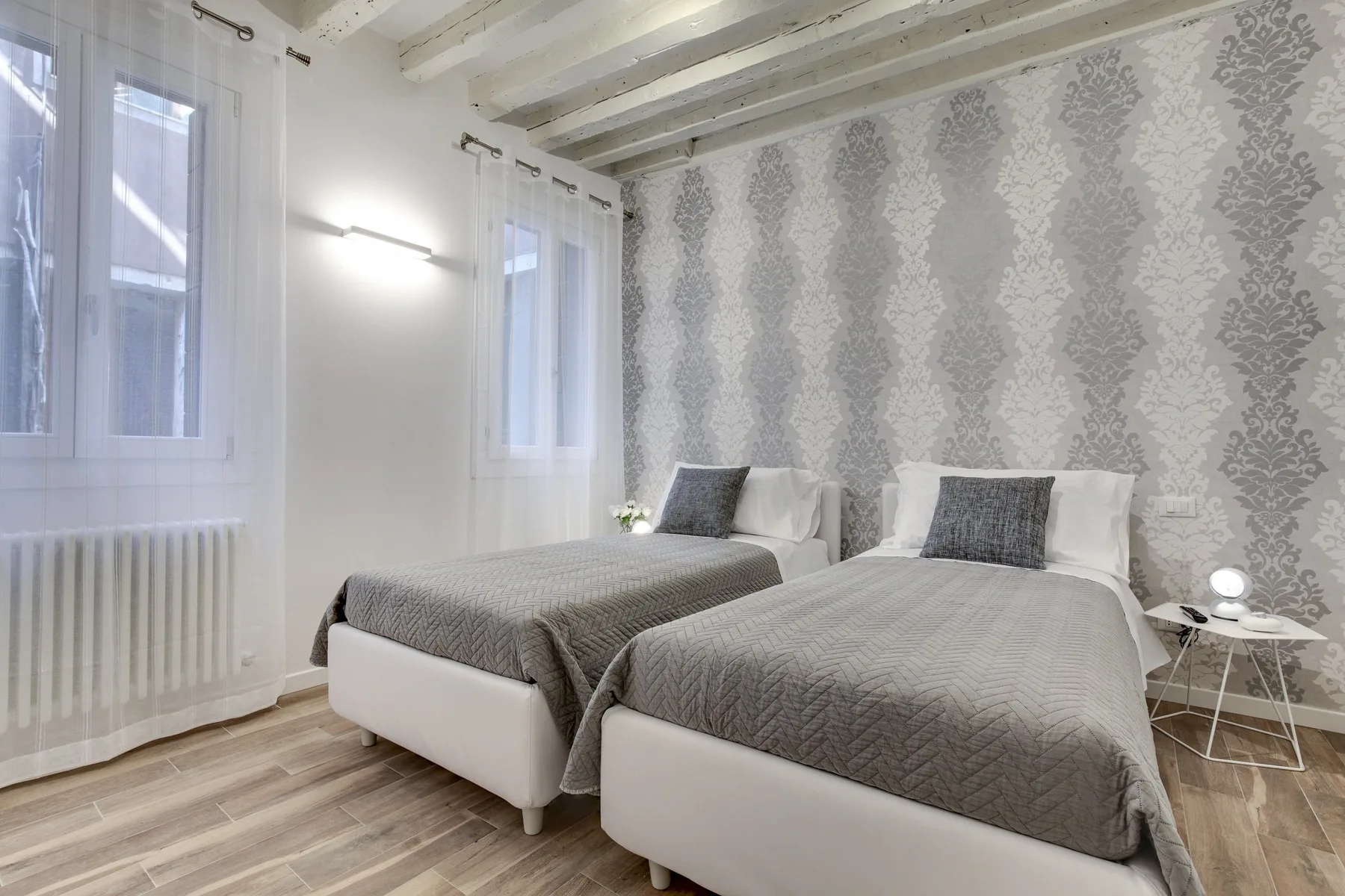 Un dormitorio con dos camas cubiertas de ropa de cama gris junto a ventanas altas.