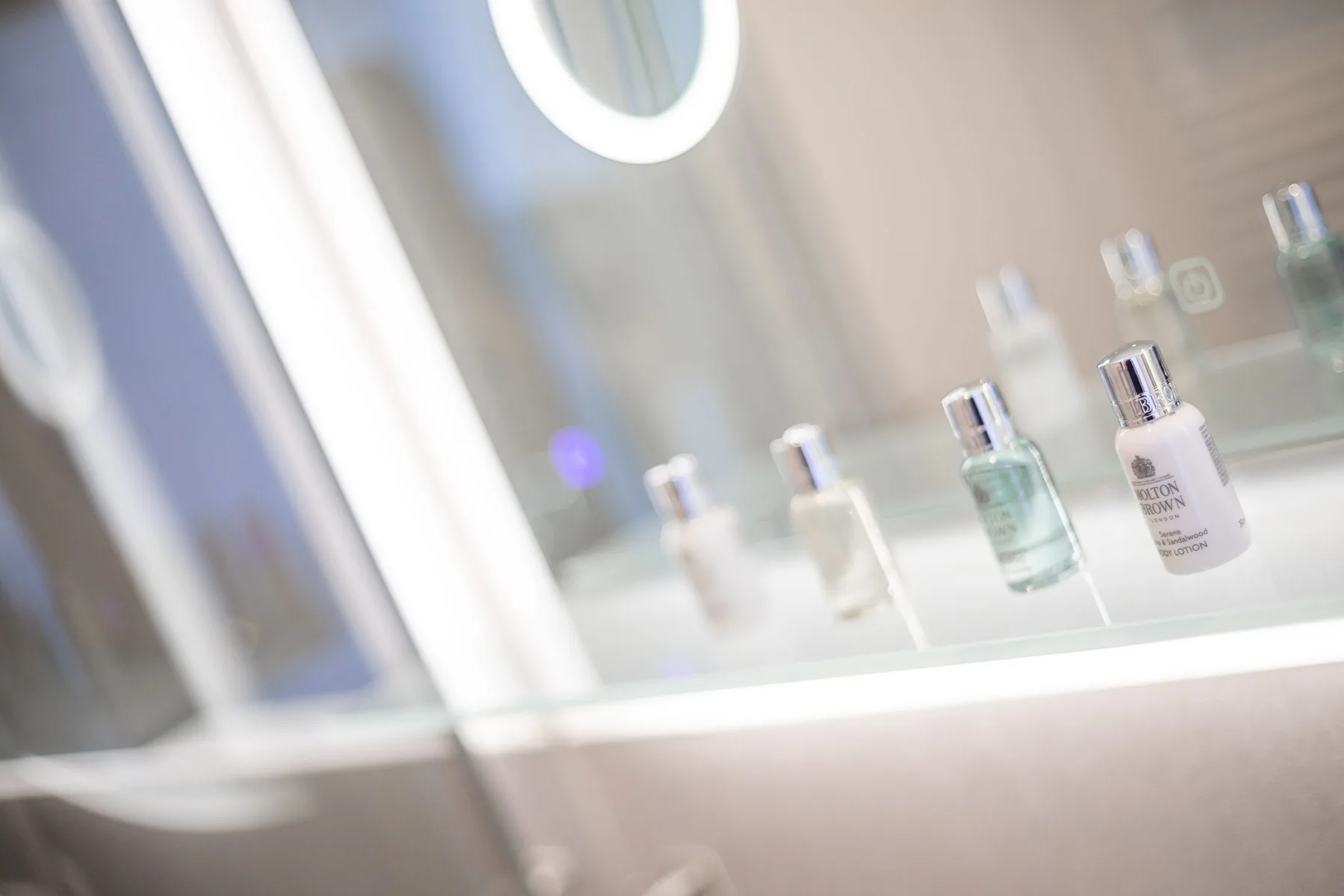 A bathroom vanity displays several small bottles beneath a lit mirror.