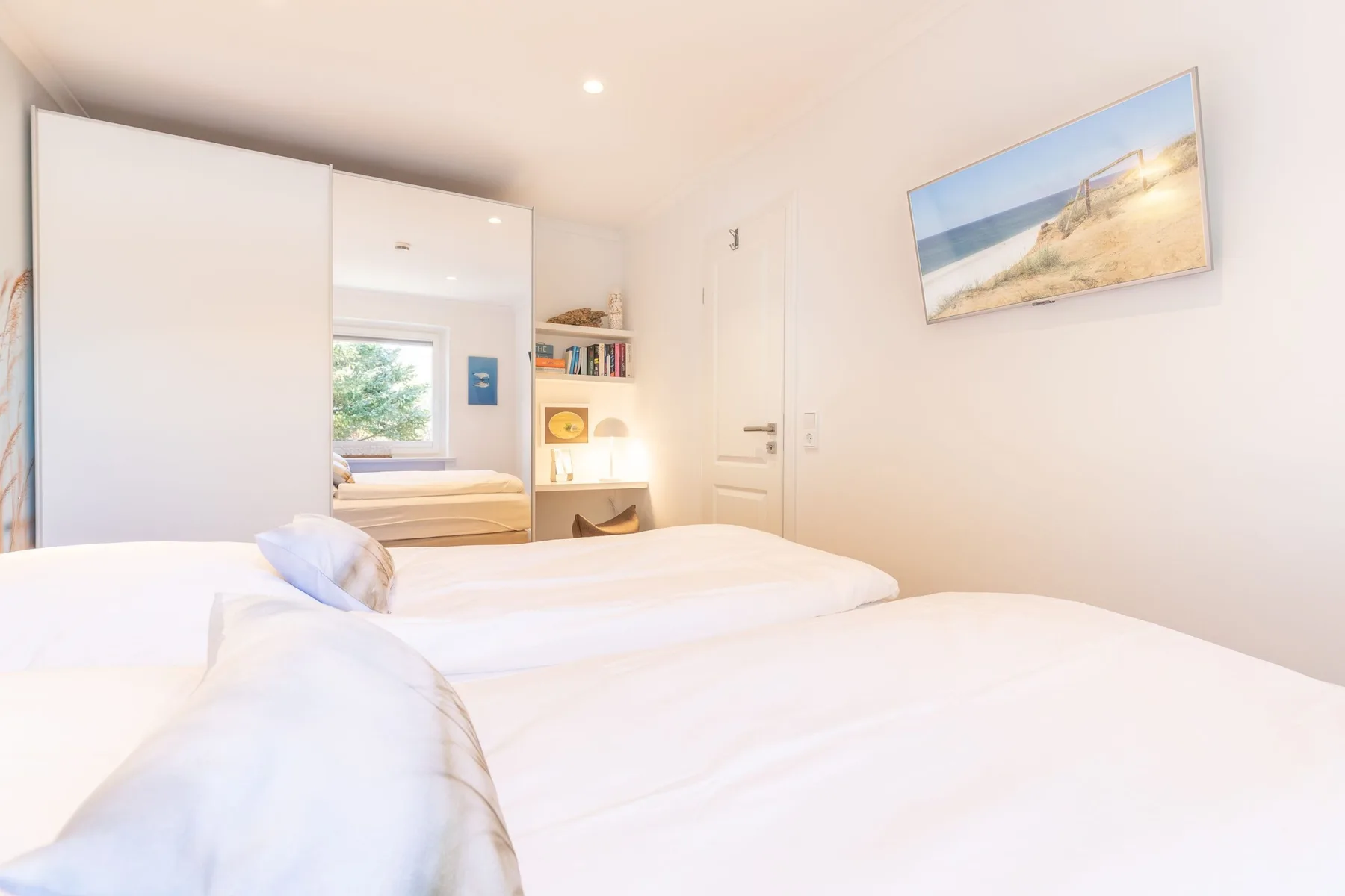 A bright bedroom with white bedding and built-in shelving beside the window.