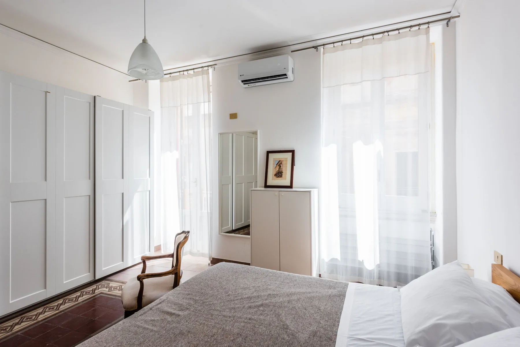 A bright bedroom with a bed, tall windows, and sheer curtains.