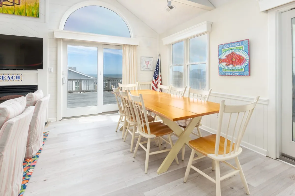 A bright dining area with a long wooden table beside large windows.