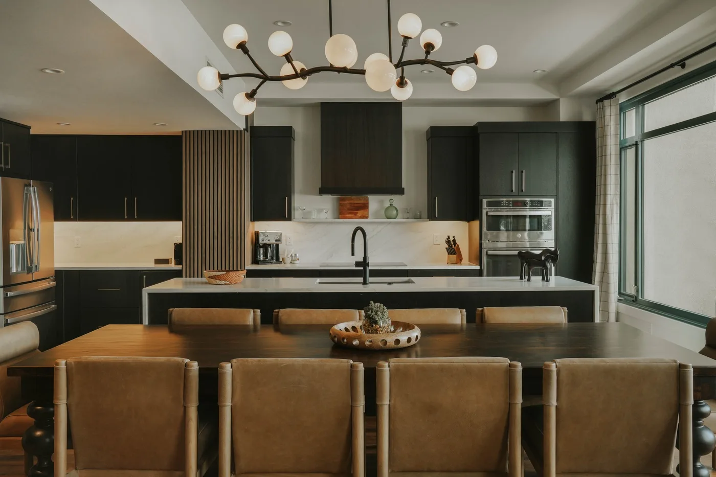 A modern kitchen opens onto a dining area with a long table and upholstered seating.