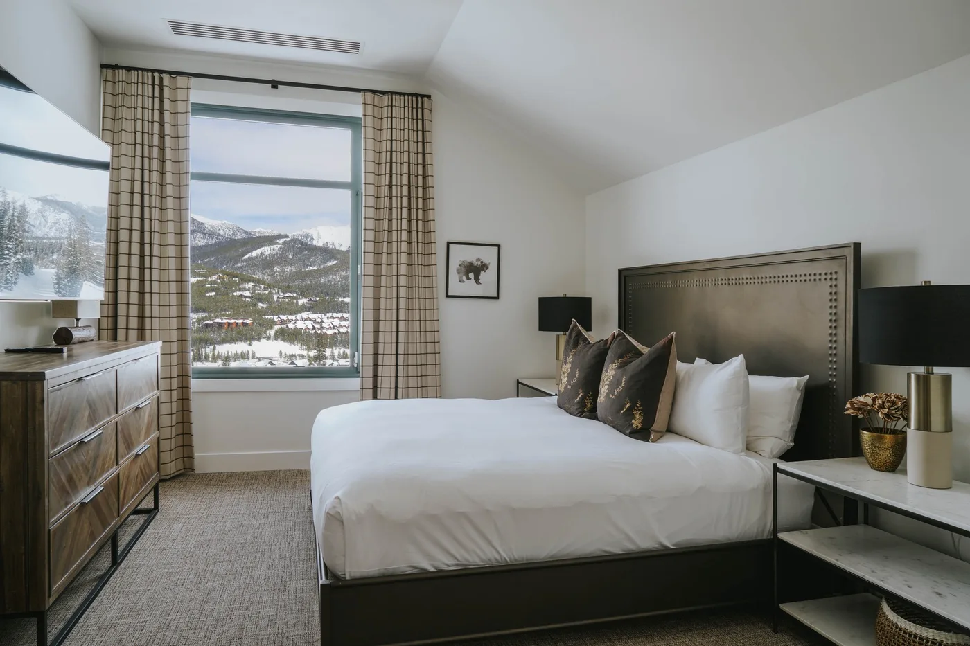 A bedroom with a neatly made bed beside a large window overlooking a snowy mountain landscape.