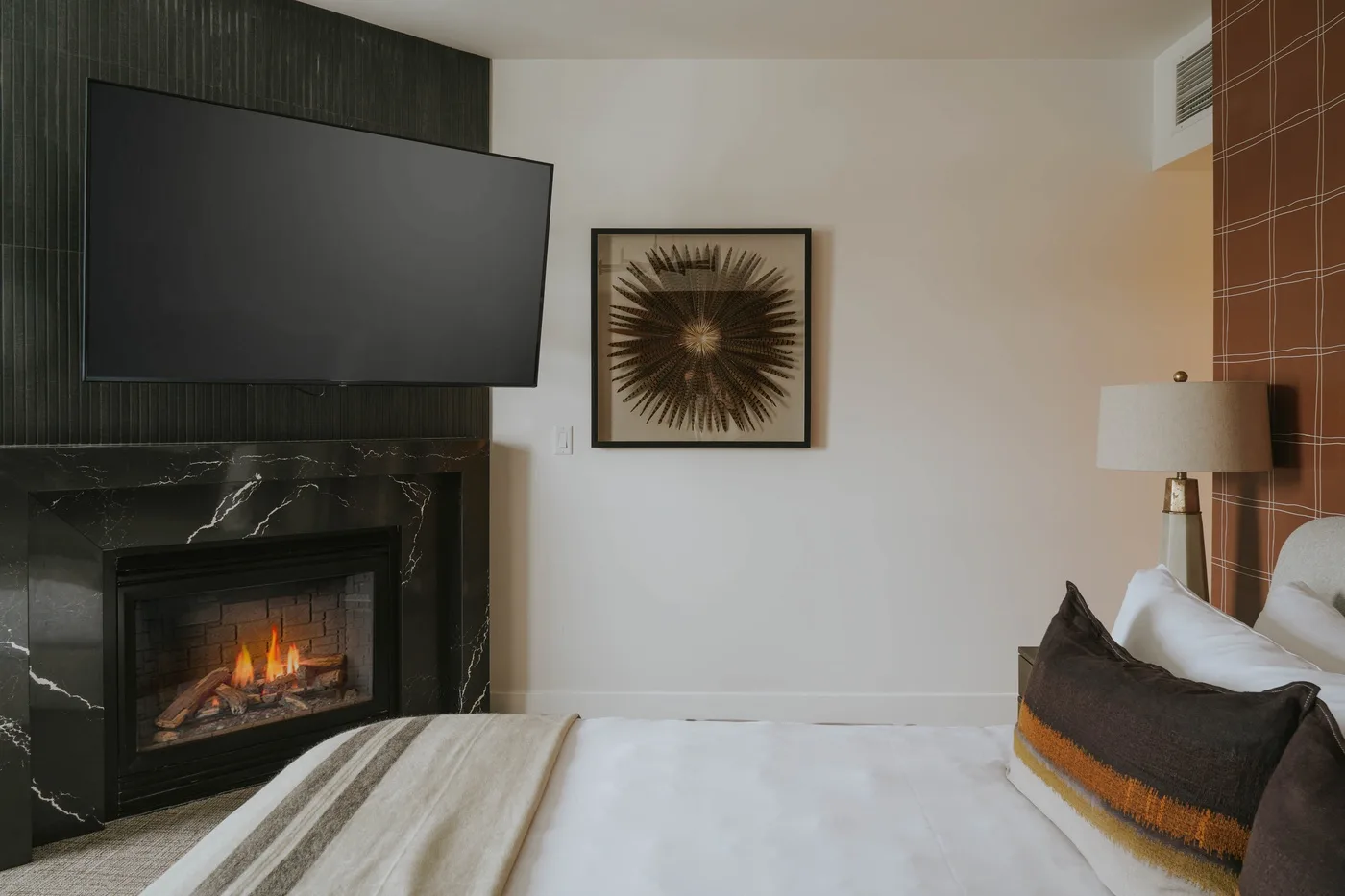 A bedroom with a neatly made bed, framed artwork, and a fireplace beneath a television.