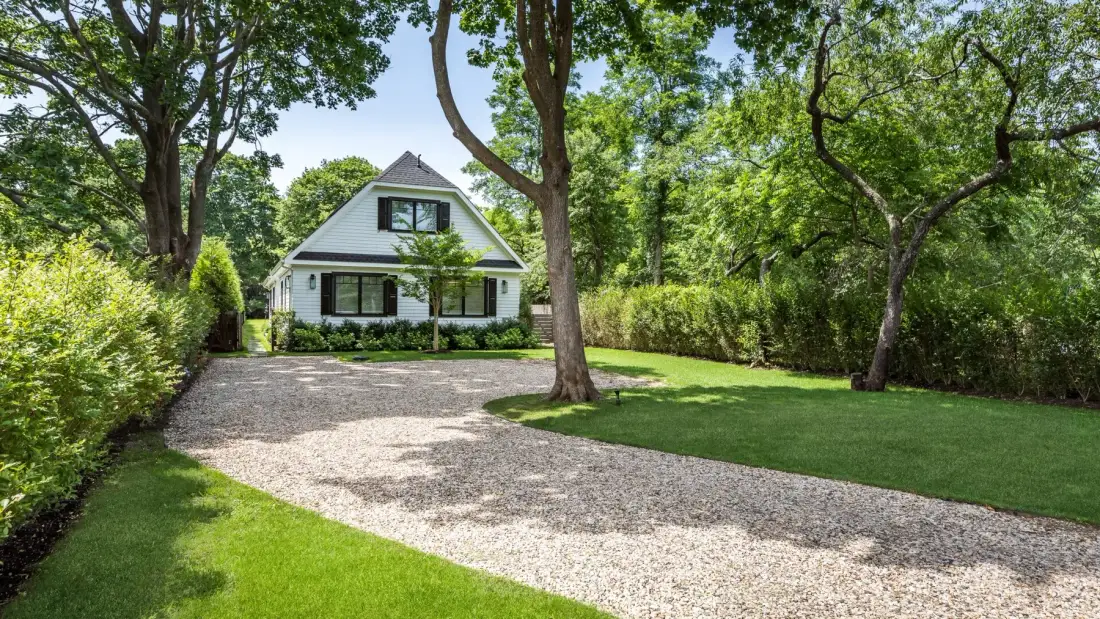 A house stands beside a gravel driveway, surrounded by trees and lawn.
