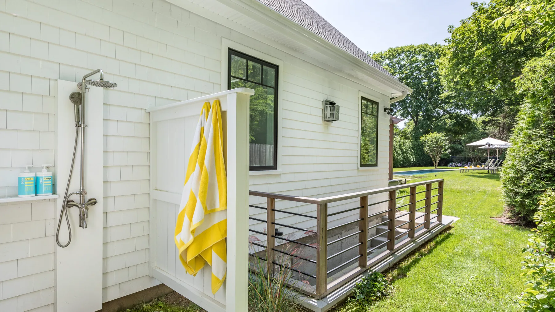 An outdoor shower beside a white house, with a wooden deck and green lawn.