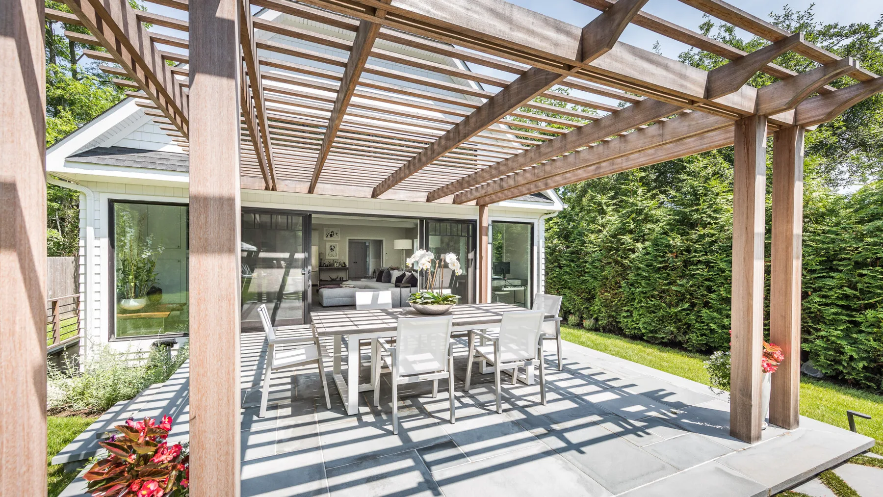 An outdoor dining area sits beneath a wooden pergola beside a glass-walled house.