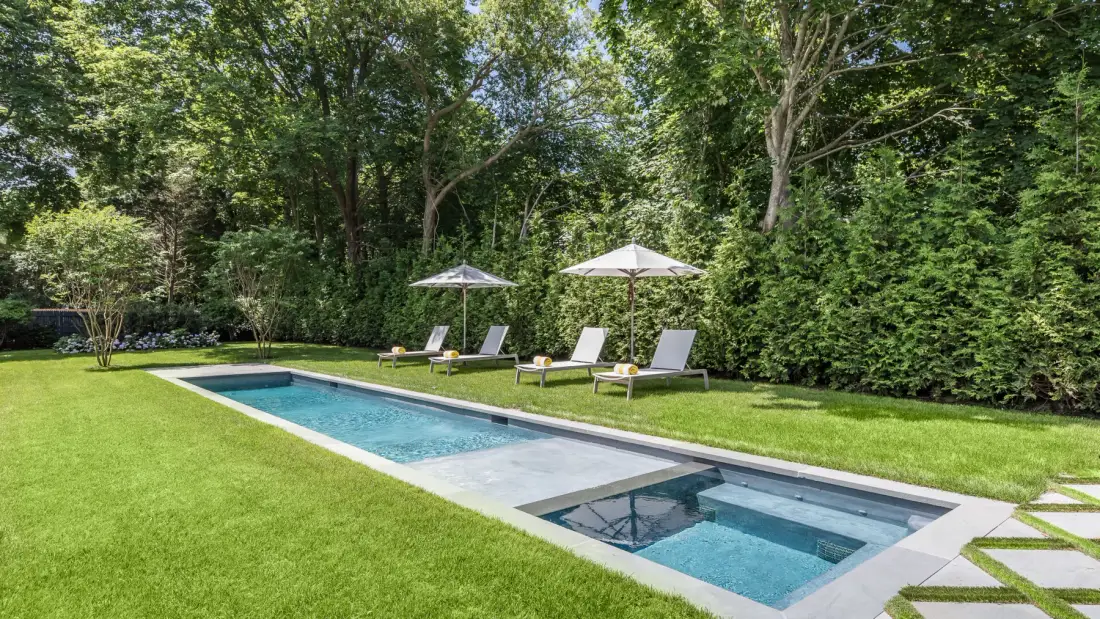 An outdoor swimming pool sits beside a lawn, with shaded seating nearby.