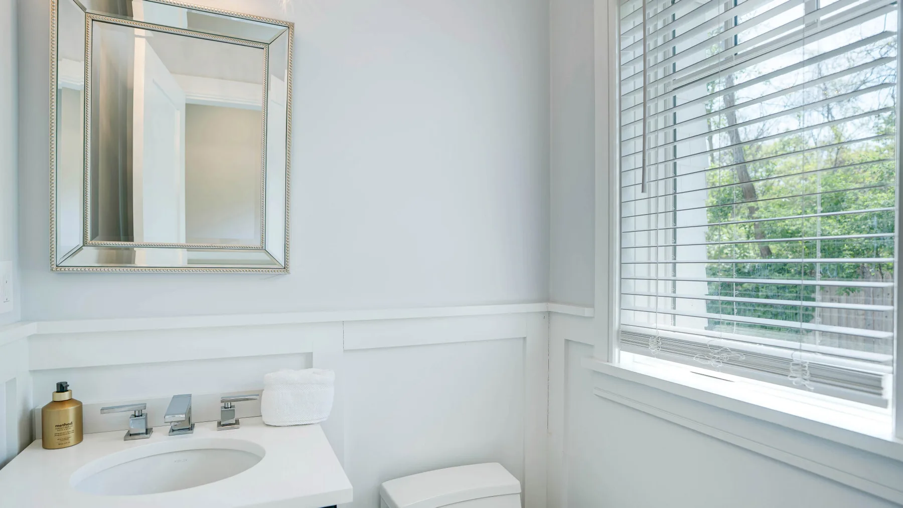 A bright bathroom with a sink, mirror, and window with blinds.