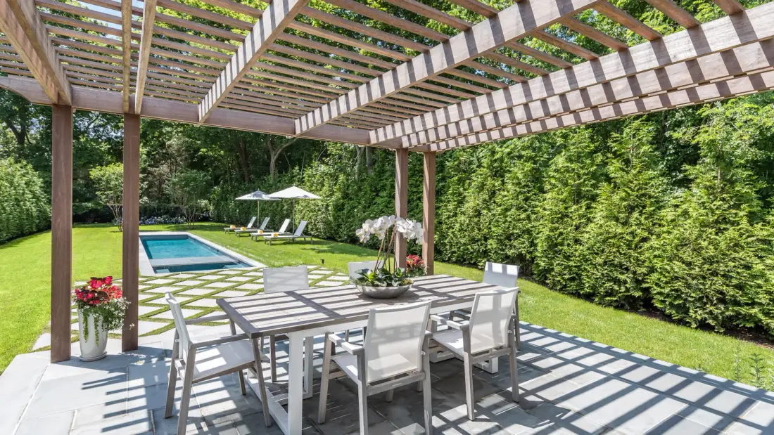 A covered patio with outdoor dining furniture overlooks a lawn and swimming pool.