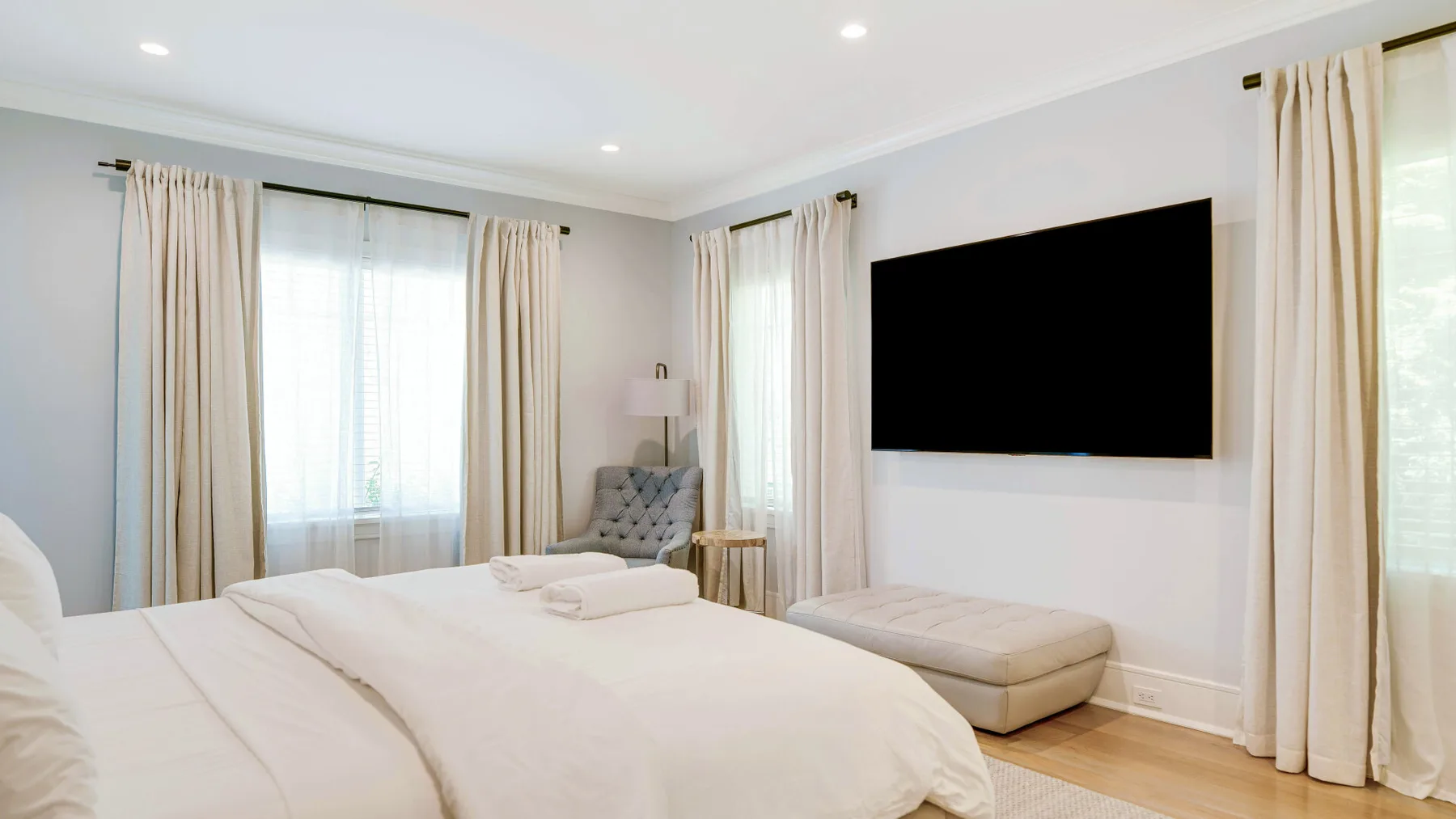 A bright bedroom with a bed, a wall-mounted television, and large curtained windows.