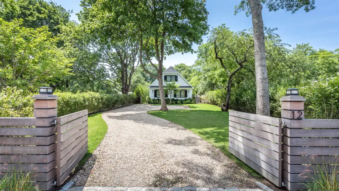 A gravel driveway leads through an open wooden gate toward a house among trees.