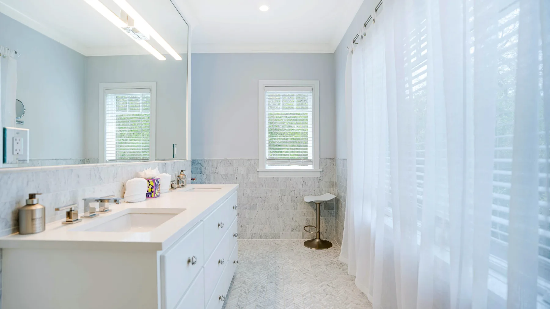 A bright bathroom with a long vanity, tiled walls, and sheer curtains.