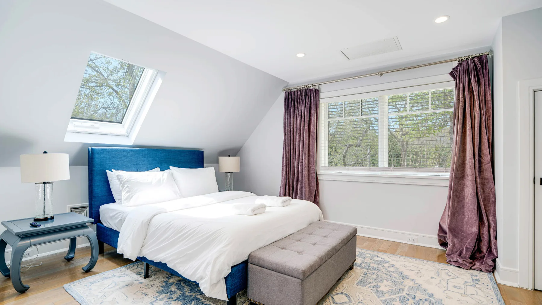 A bright bedroom with a blue bed, bedside tables, and a wide window.