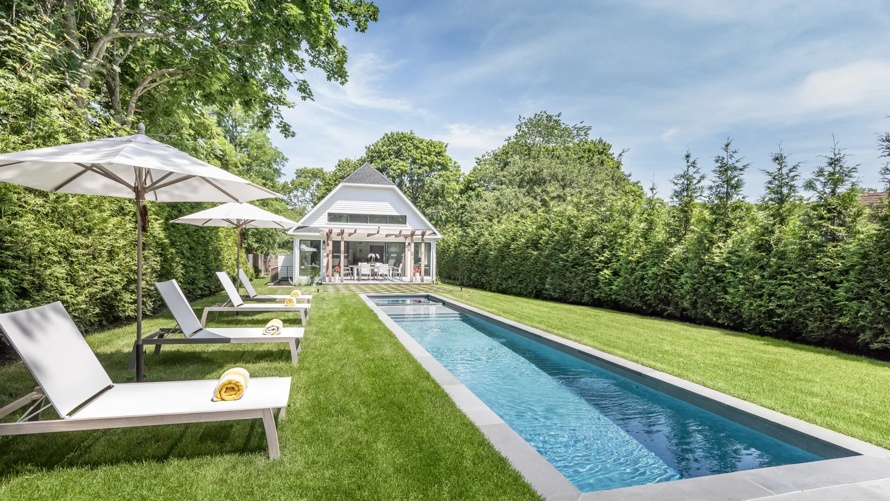 A long outdoor swimming pool runs beside a grassy lawn and lounge seating.