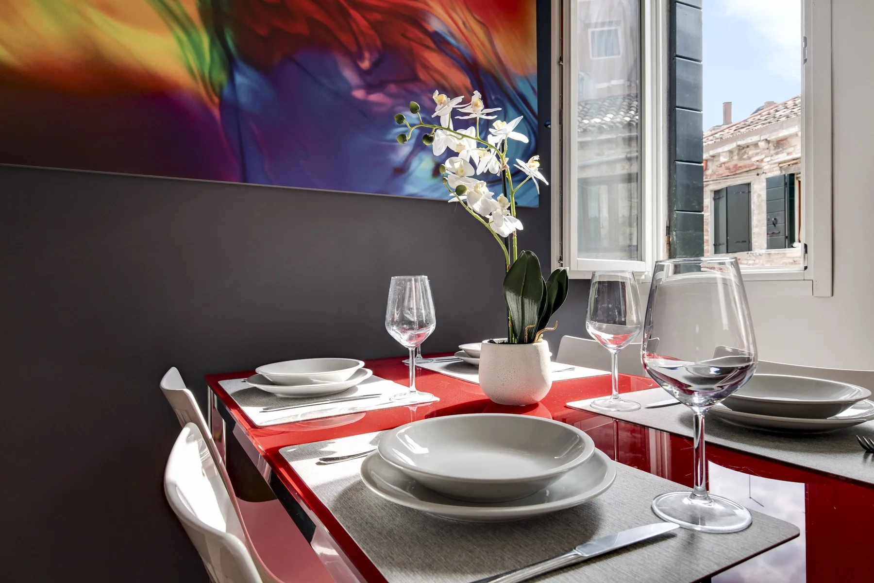 A dining table set with plates and glasses sits beside a bright window.